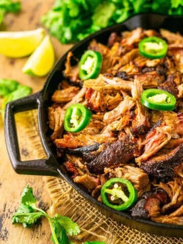 The smoked carnitas in a black cast-iron skillet with jalapeños scattered around and lime slices and cilantro in the background.