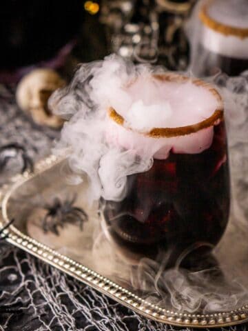 Two glasses of Halloween sangria with mist coming out of them on a silver tray with twinkle lights and a decorative skull in the background.