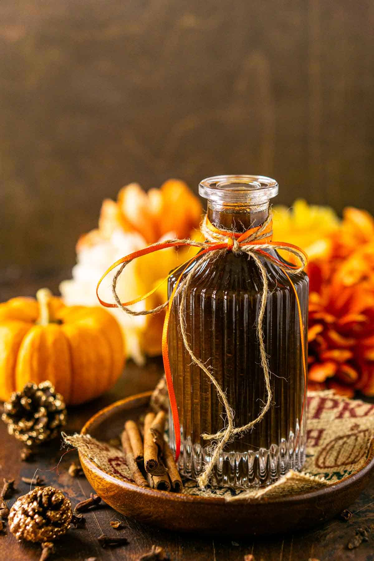 A straight-on view of a bottle of pumpkin spice simple syrup against a brown wooden surface with flowers in the background.