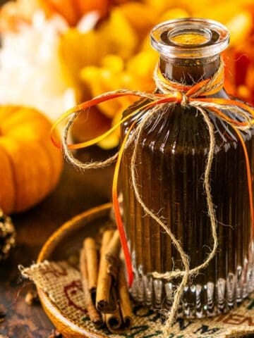 A bottle of pumpkin spice syrup on a brown wooden plate with fall flowers in the background and whole cloves scattered around it.