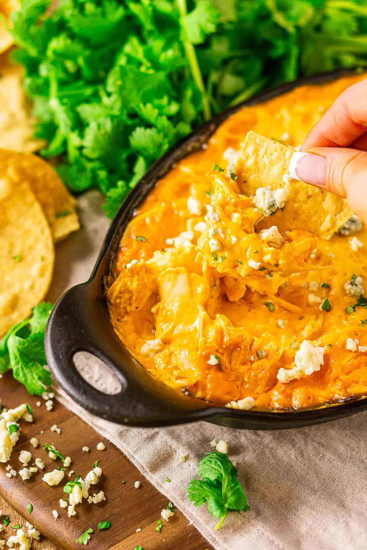 A hand dipping a tortilla chip into the smoked Buffalo chicken dip on a wooden plate with a neutral-colored napkin folder underneath the baking dish.