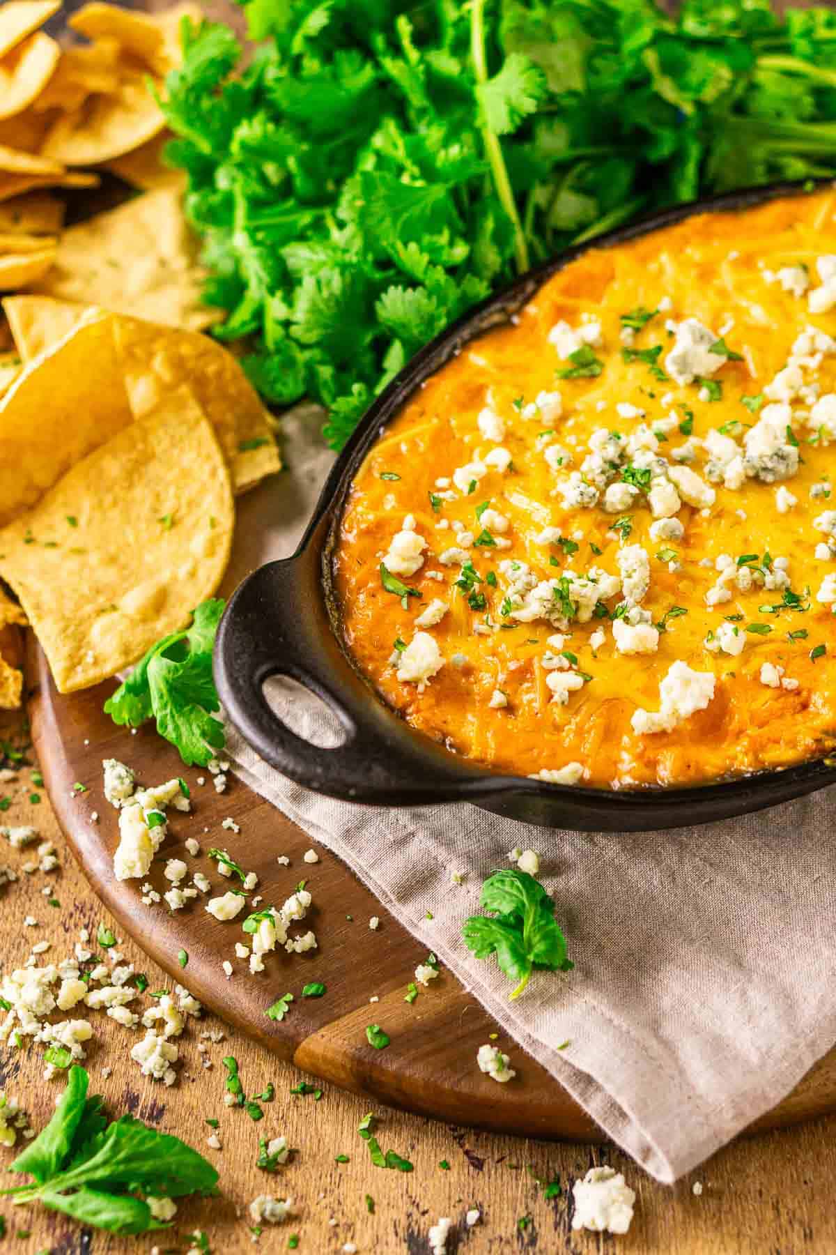 A small cast-iron baking dish filled with the smoked Buffalo chicken dip on a wooden plate with blue cheese crumbles around it and a bundle of cilantro in the background.