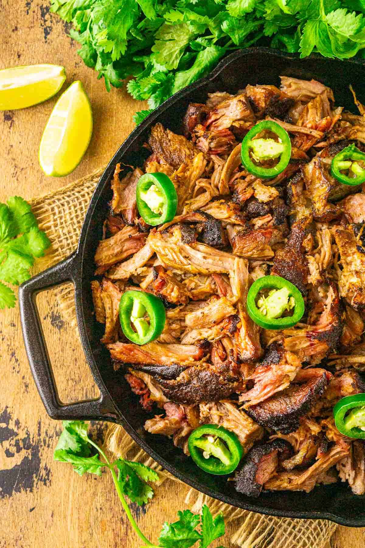 An aerial view of the smoked carnitas in a black cast-iron skillet with a bundle of fresh cilantro on top.