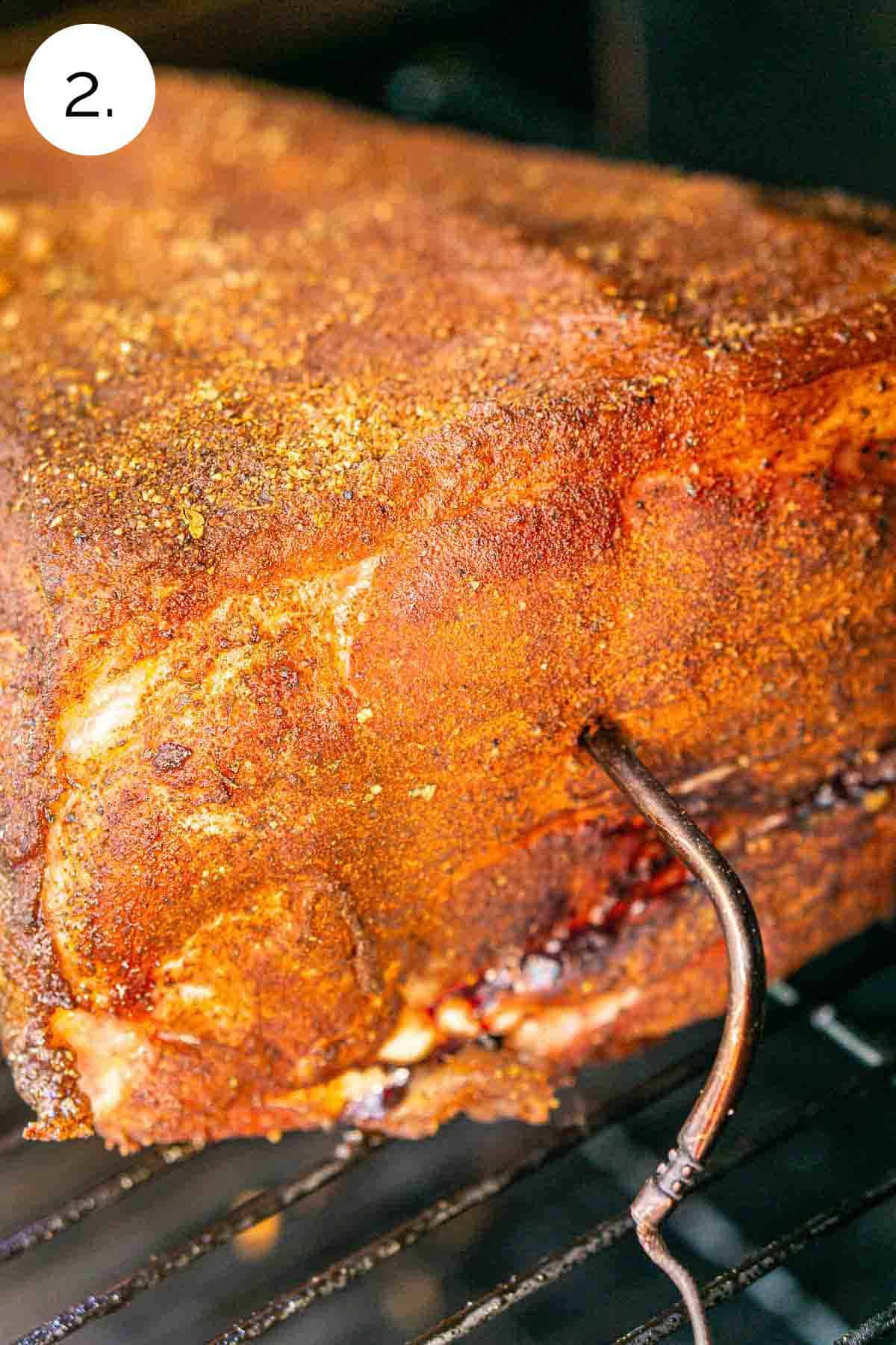 The seasoned pork butt on the grates of the smoker with a leave-in meat thermometer inserted in the center of the roast.