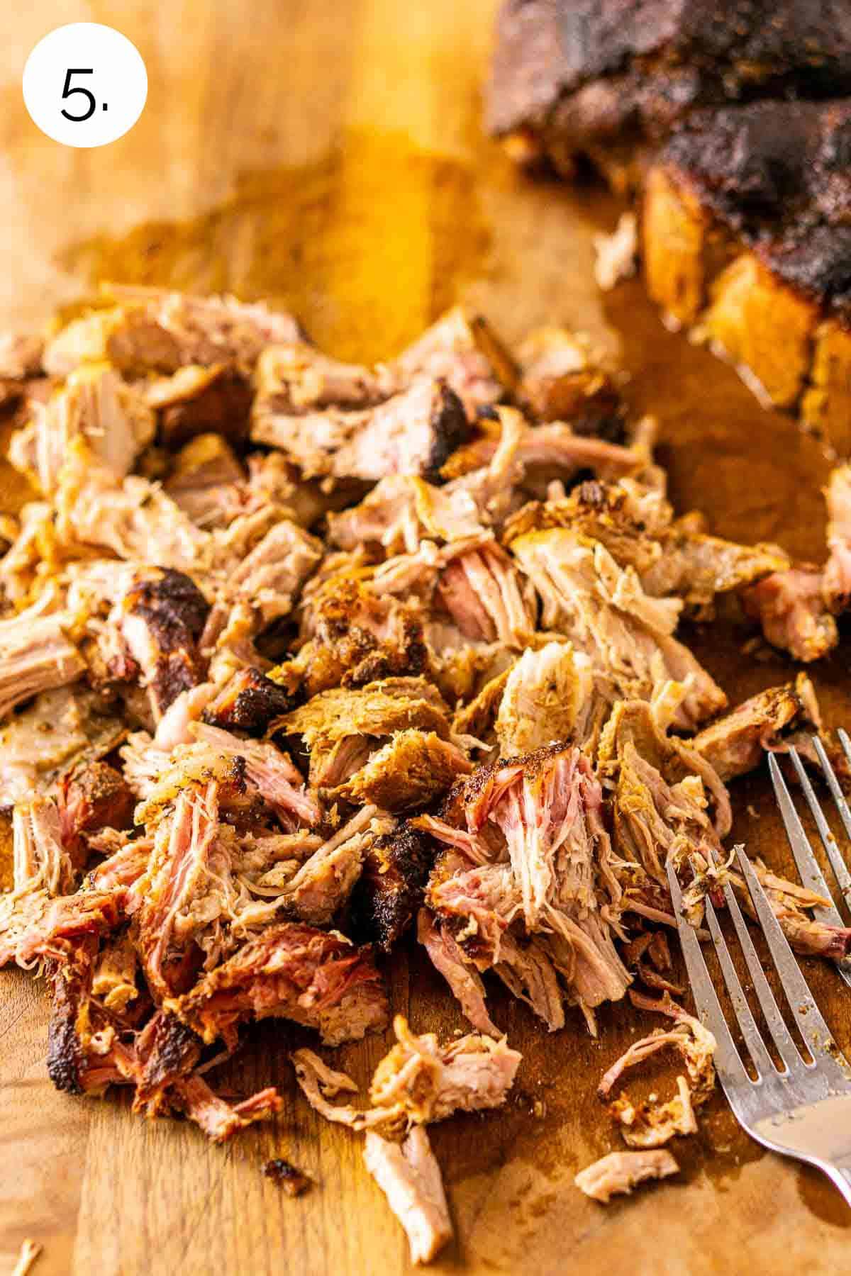 The carnitas on a brown cutting board with two forks to the side after the meat has been shredded.