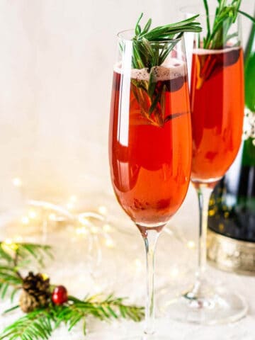 Two cranberry mimosa drinks on a wintry white surface with a bottle of Champagne and Christmas decor surrounding them.