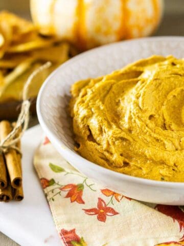 A bowl of spicy pumpkin hummus on a fall-themed napkin with a bundle of cinnamon to the left and a real pumpkin in the background.