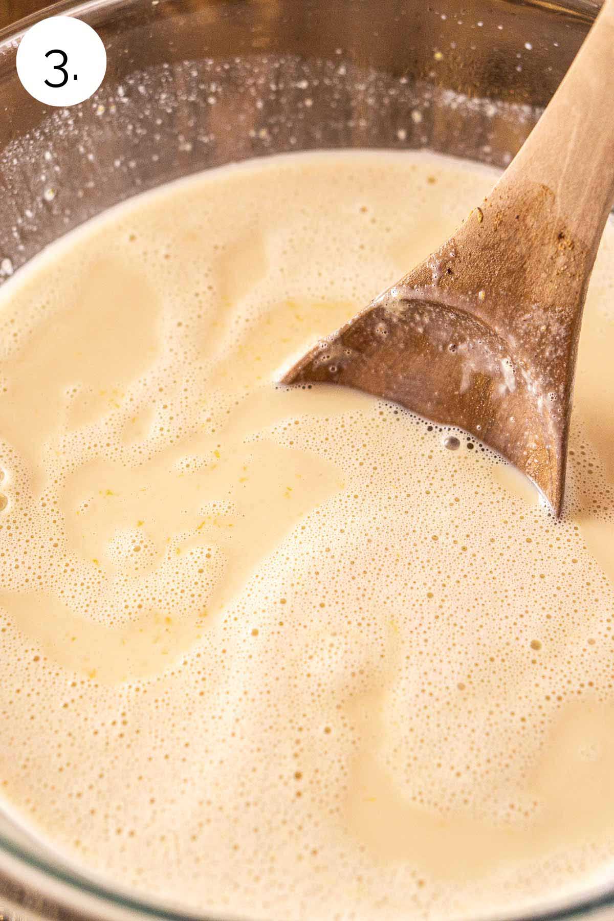 A wooden spoon stirring the bourbon into the eggnog mixture in a large glass mixing bowl.