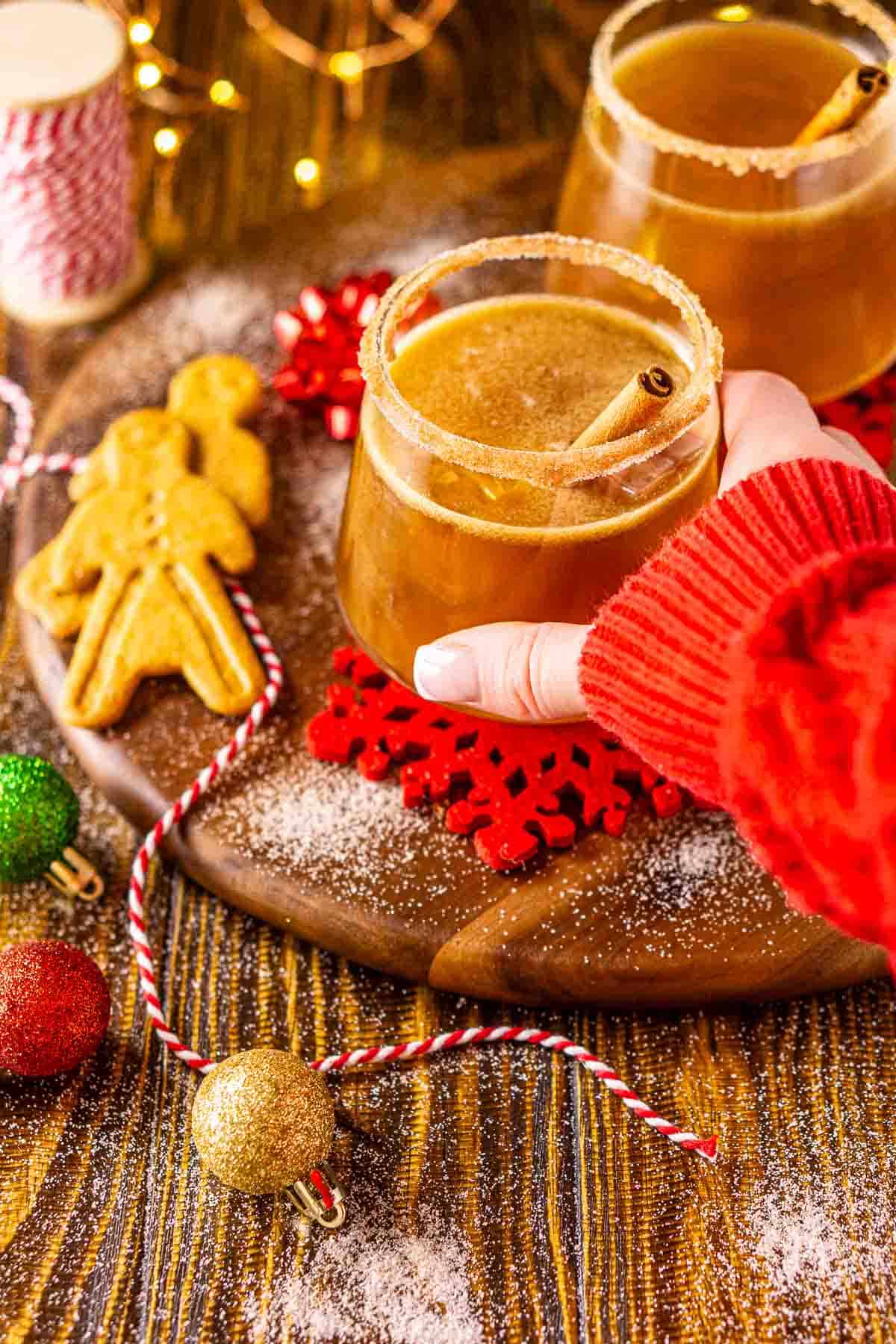 An arm in a red Christmas sweater reaching out to pick up the gingerbread margarita on a wooden plate with holiday decor around it.