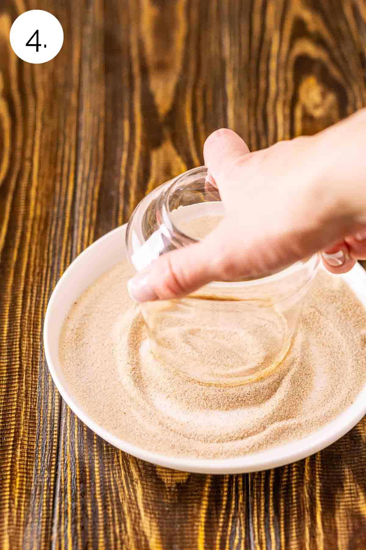 A hand dipping a glass into a mixture of cinnamon and sugar on a small white plate.