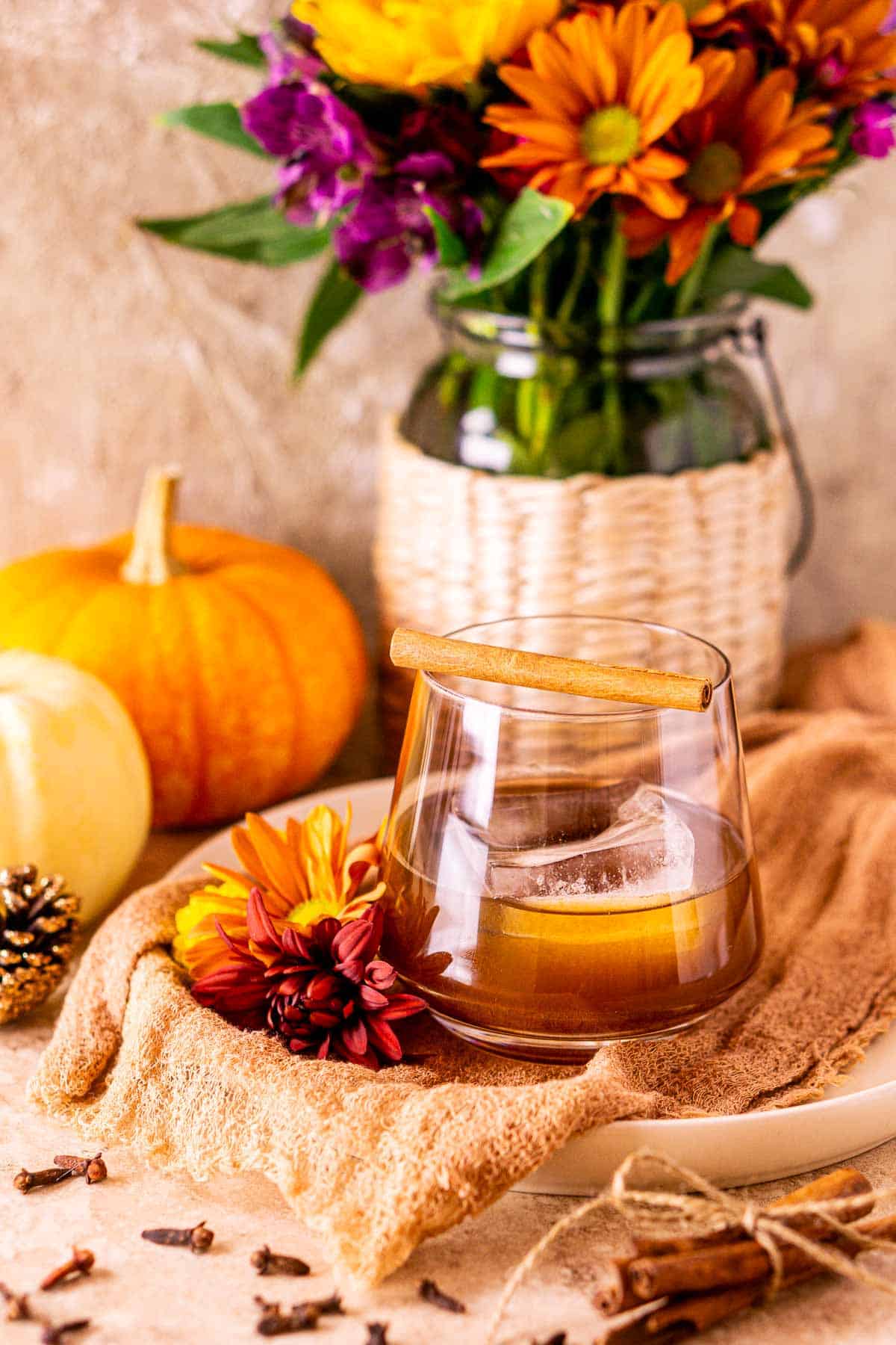 A pumpkin old fashioned on a white plate with fall-colored cloth and colorful flowers on the side of the glass.