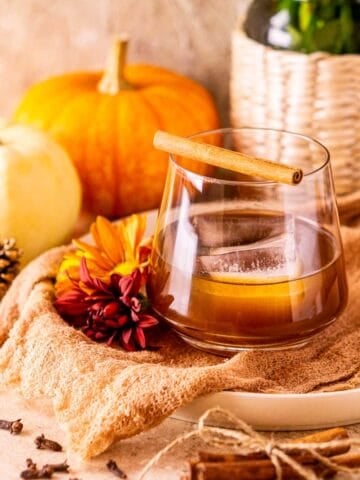 A spiced pumpkin old fashioned on a cream-colored cloth with spices around it and a vase of fall decor in the background.