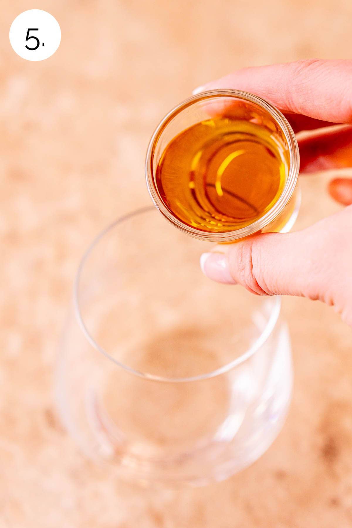 A hand pouring a shot of bourbon into an old fashioned glass on a cream-colored tile surface.