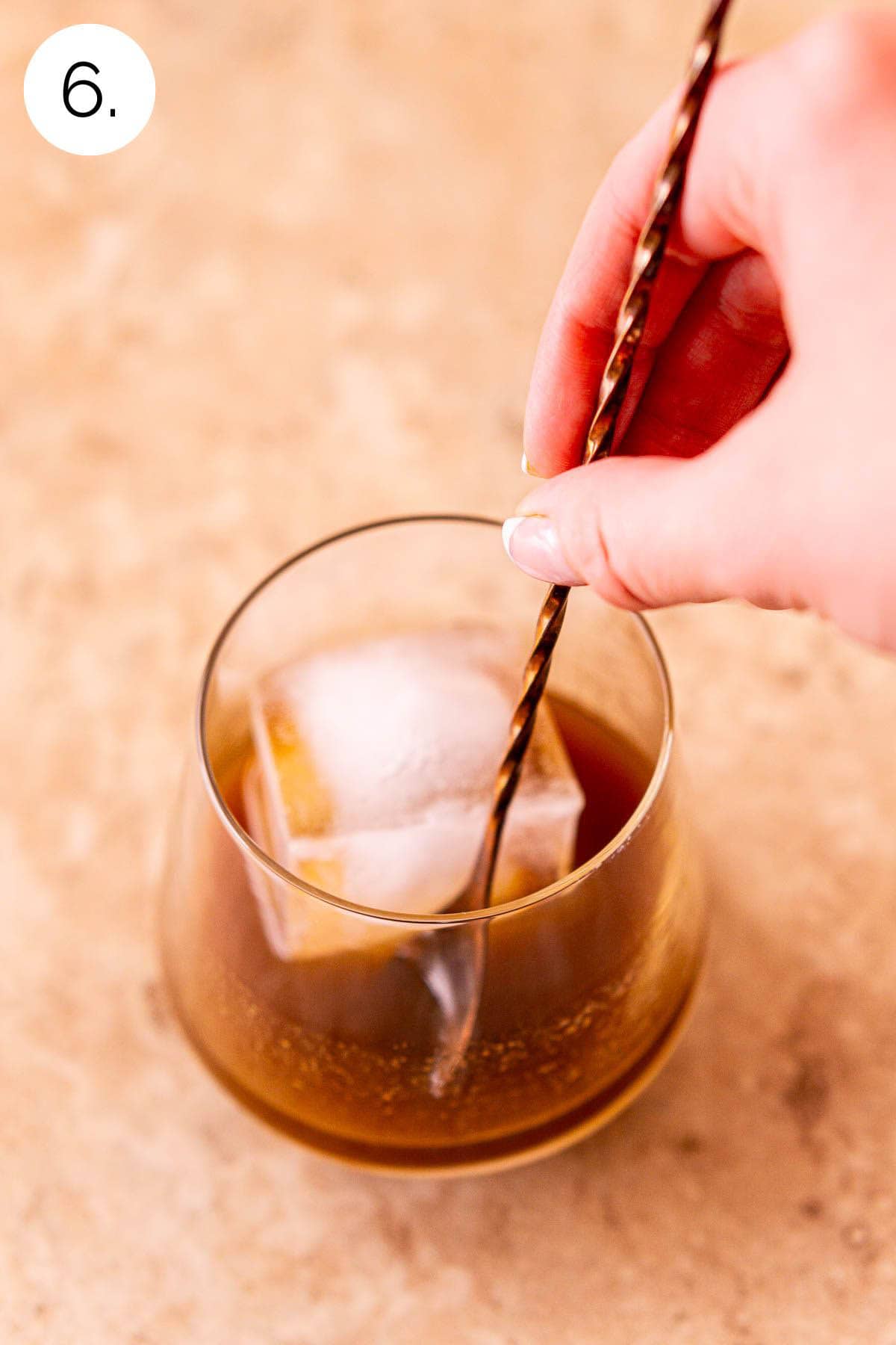 A hand stirring the old fashioned and large ice cube with a copper bar spoon.