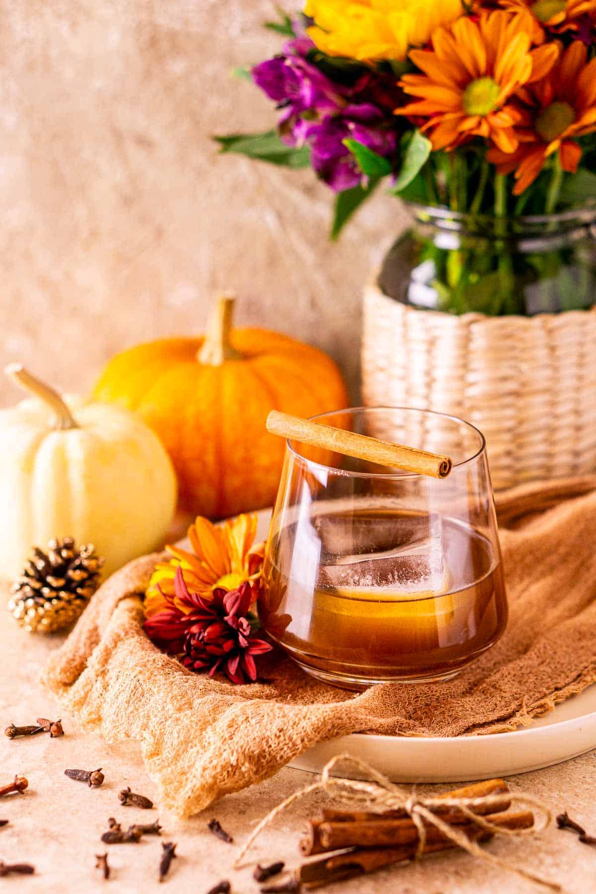 A spiced pumpkin old fashioned on a cream-colored cloth with spices around it and a vase of fall flowers in the background.