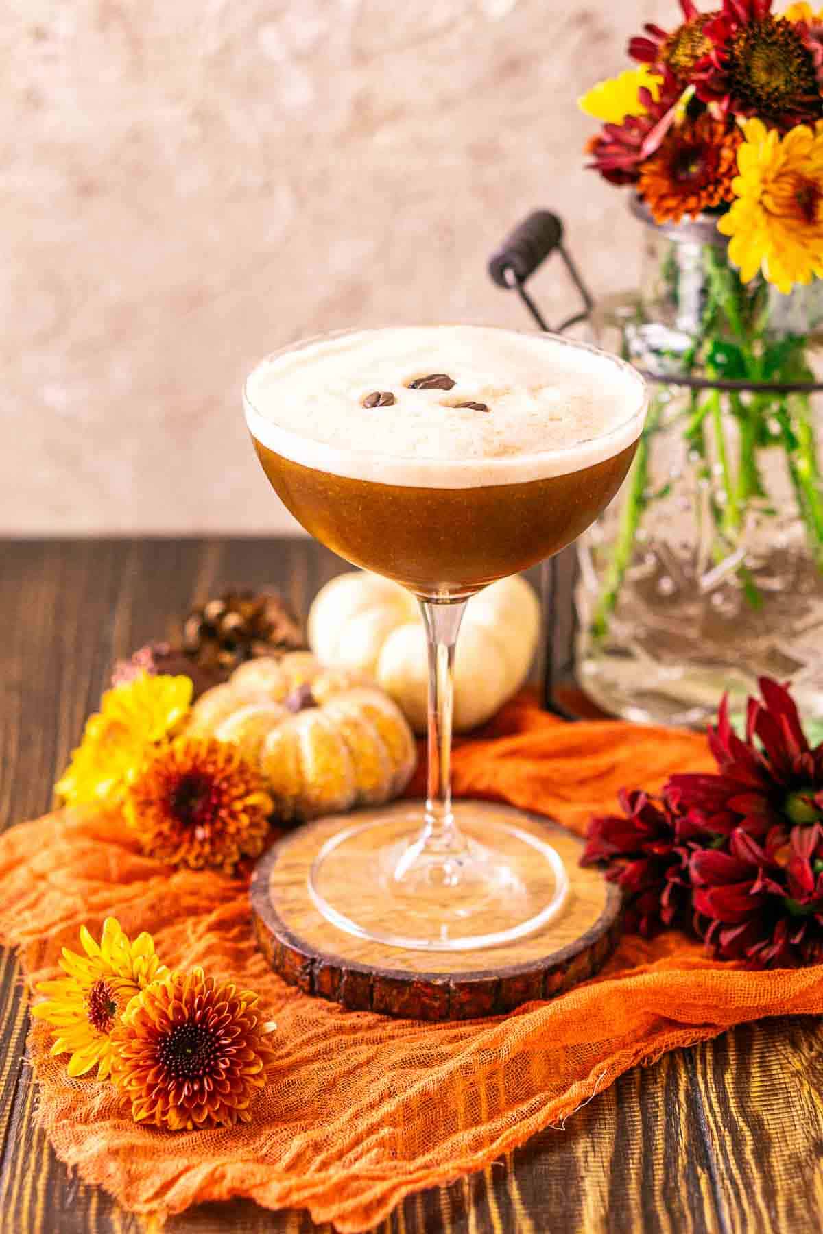 A pumpkin spice espresso martini on a wooden coaster with fall-colored flowers around it and a vase to the right.