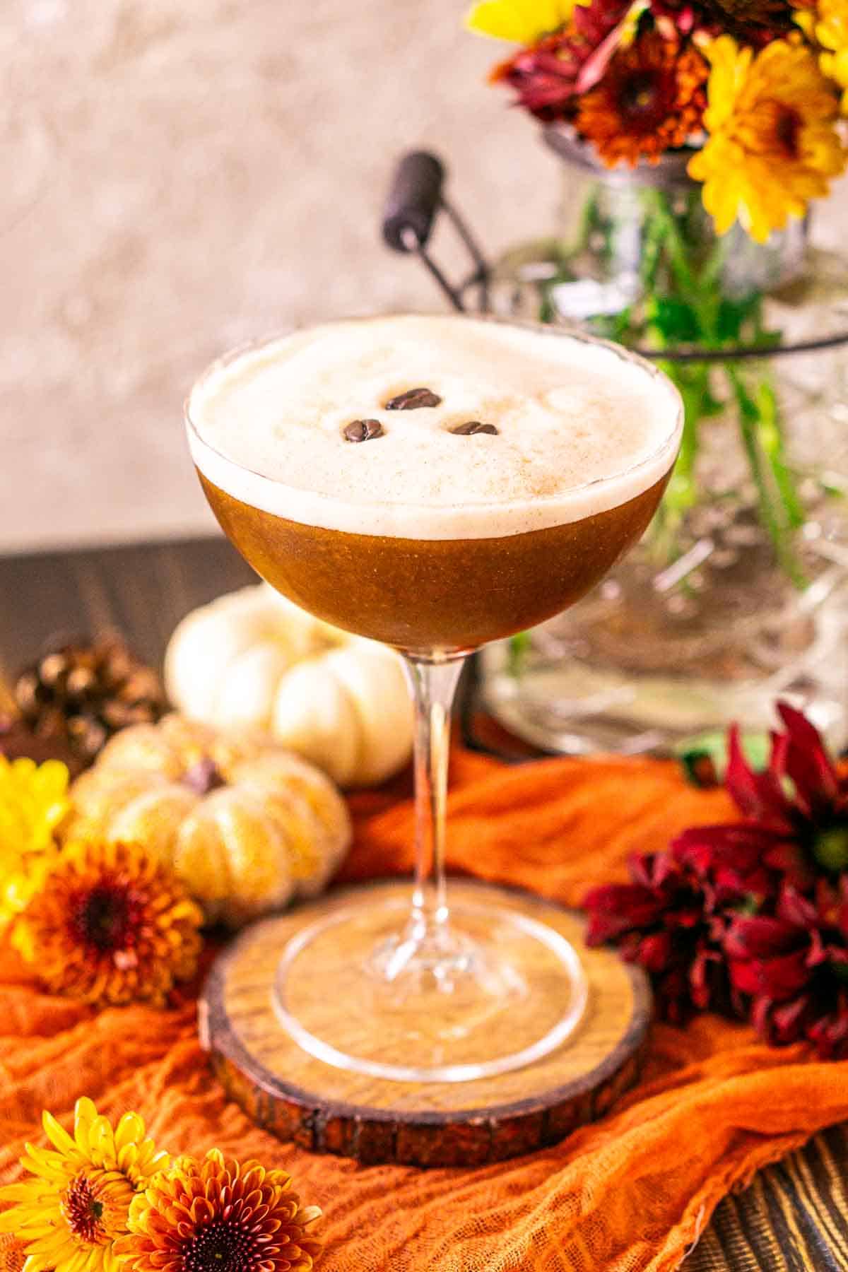A close-up view of the pumpkin spice espresso martini on a wooden coaster with orange cloth underneath and colorful flowers around it.