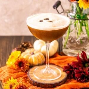 A pumpkin spice espresso martini on a wooden coaster with miniature pumpkins in the background and fall flowers around it.