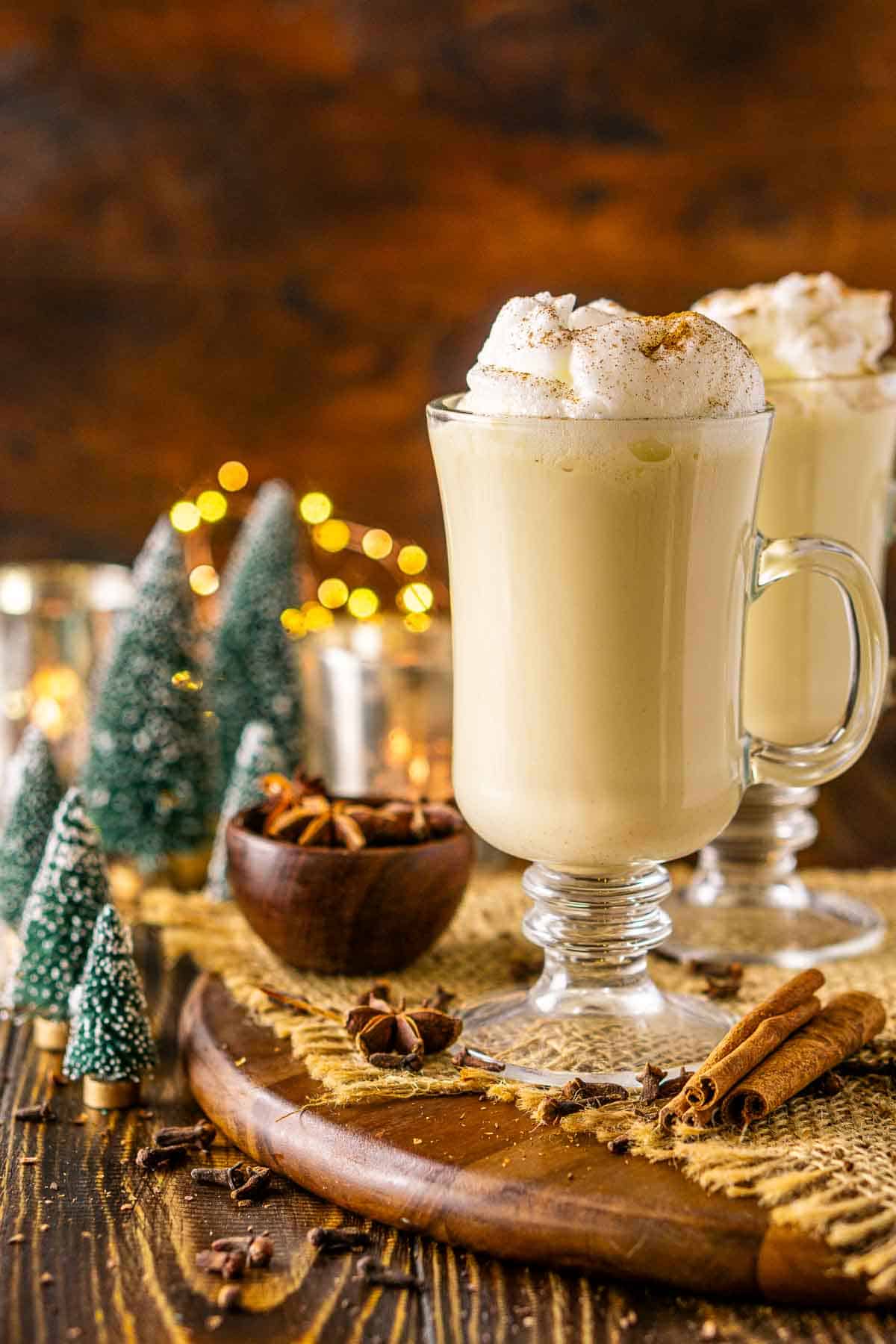 A straight-on view of the bourbon eggnog on a wooden platter with spices and Christmas decor around it against a brown background.