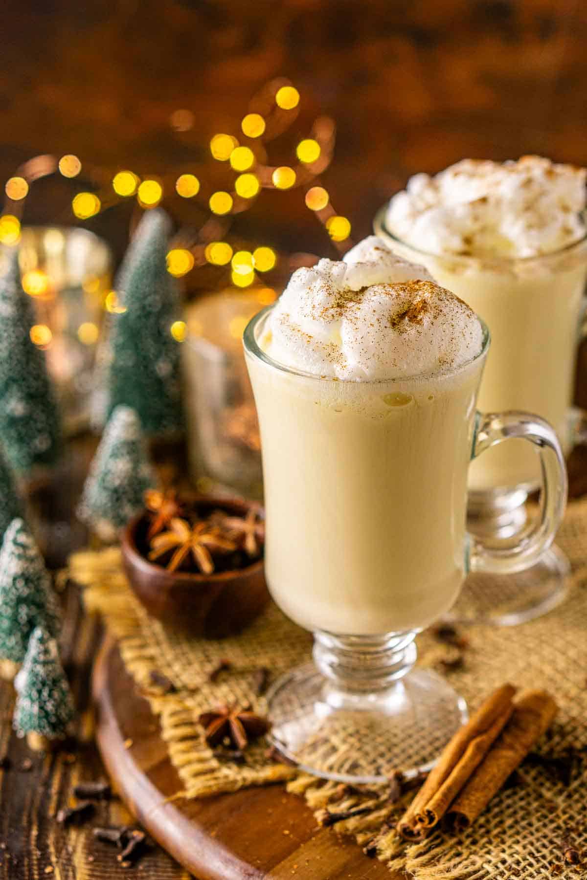 A close-up of a bourbon eggnog on a wooden platter with mini Christmas trees to the left and a bundle of cinnamon sticks to the right.