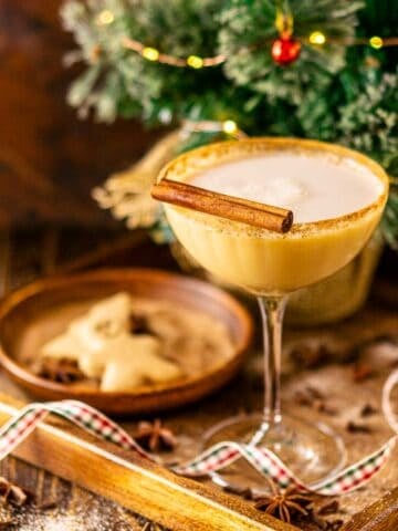 A gingerbread martini on a wooden surface with a Christmas tree in the background and holiday decor around it.