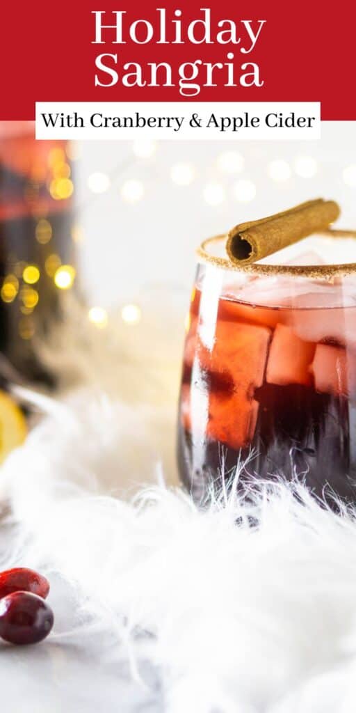 A glass of holiday sangria on a feathery white surface with festive decor around it and text overlay on top of the image.