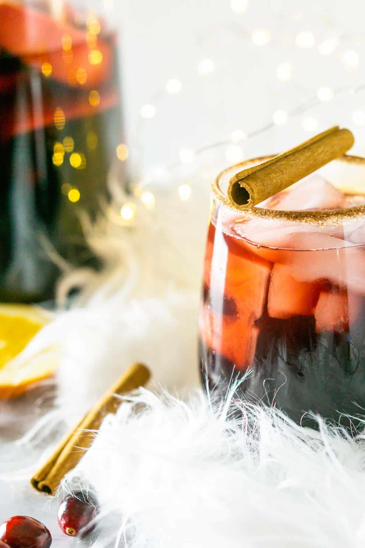A glass of holiday sangria with a cinnamon stick garnish on a white feathery piece of fabric with a pitcher and twinkling lights in the background.