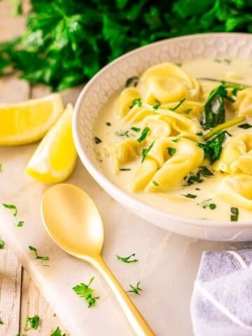A bowl of Instant Pot tortellini soup on a white marble tray with a gold spoon and lemon slices to the side and fresh parsley scattered around it.
