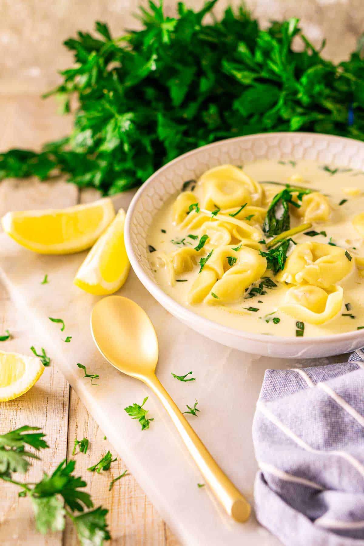 A bowl of Instant Pot tortellini soup on a white marble tray with a gold spoon and lemon slices to the side and fresh parsley scattered around it.