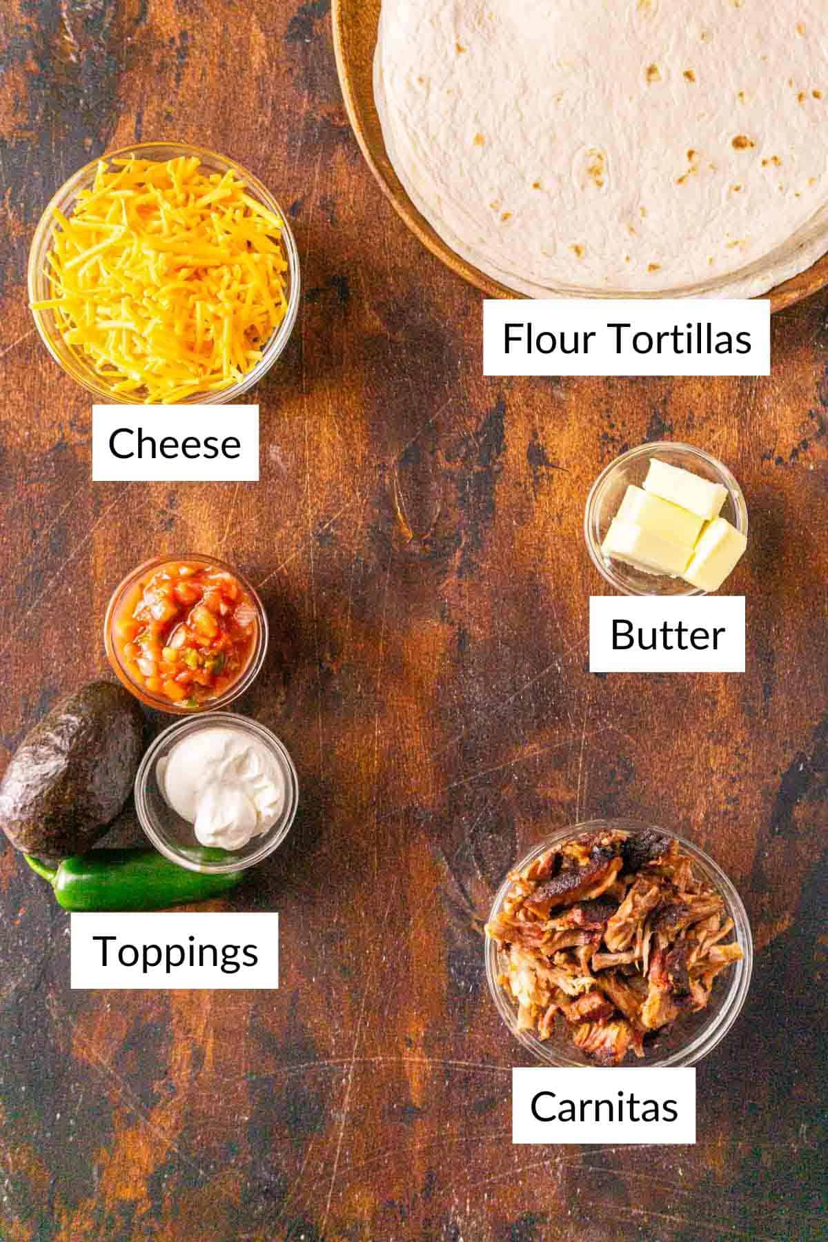The ingredients for the quesadillas on a brown and black wooden surface with white labels by the items.