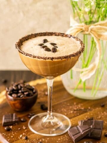 A mocha martini on a wooden tray with pieces of chocolate and coffee beans scattered around it.