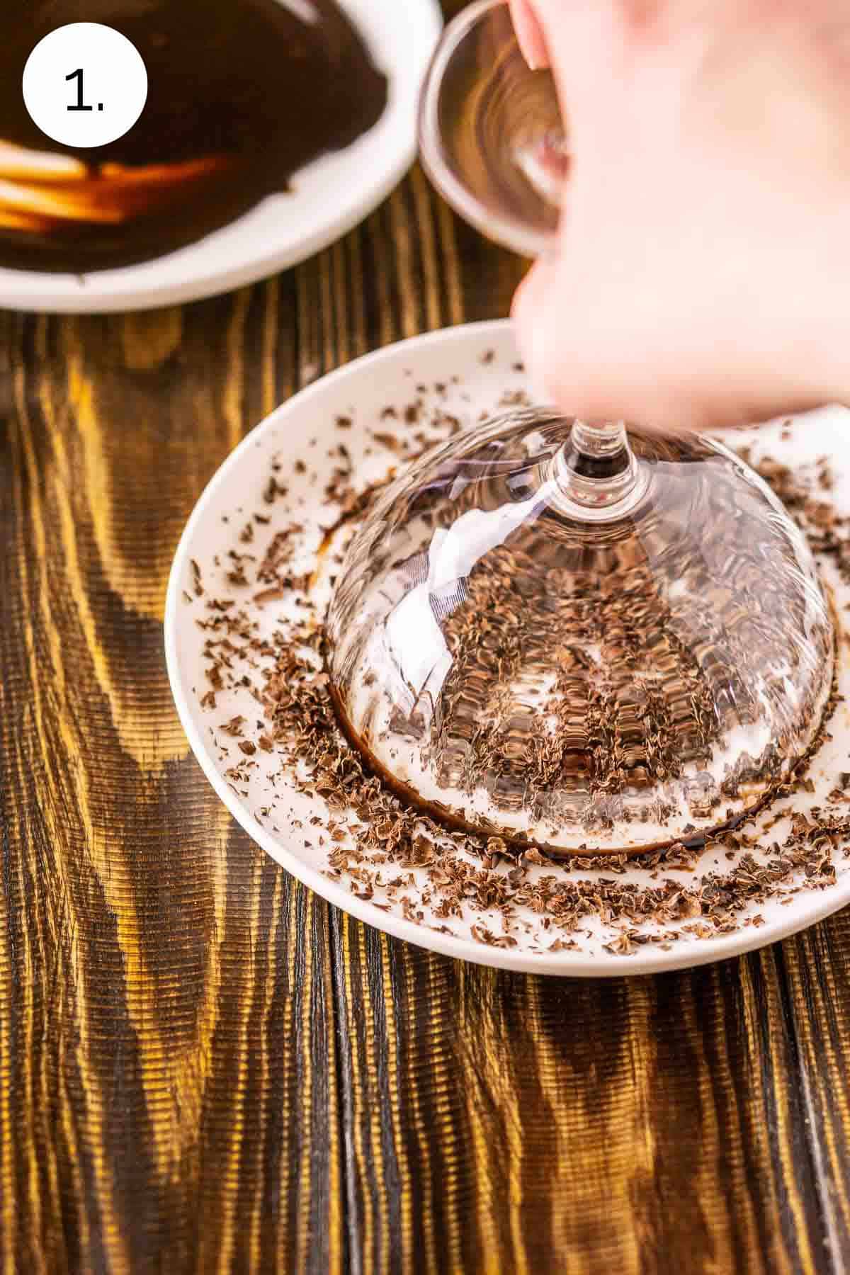 A hand dipping a martini glass covered in chocolate sauce onto a small plate with grated chocolate to garnish.