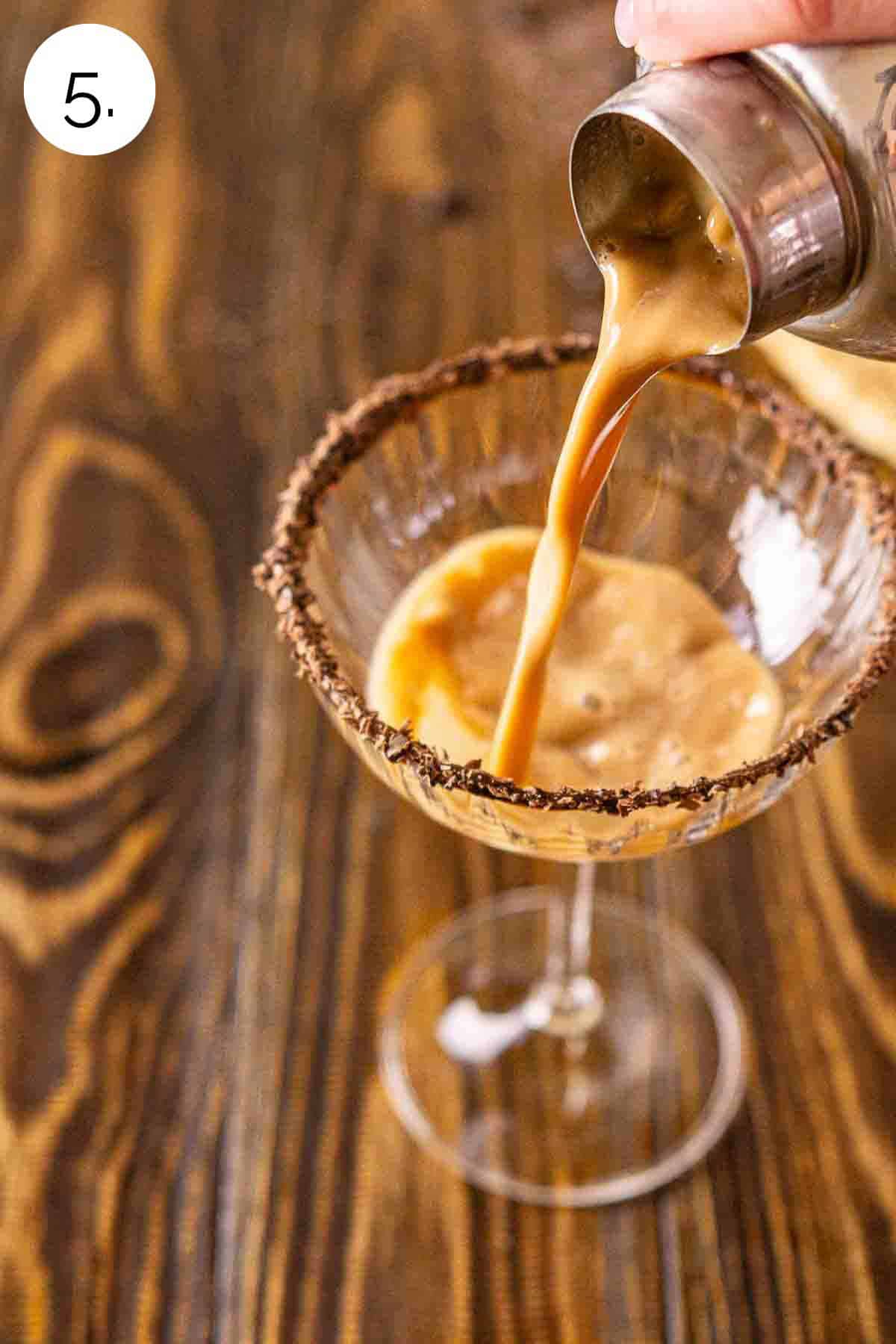 A hand straining the drink into a garnished martini glass after shaking on a brown wooden surface.