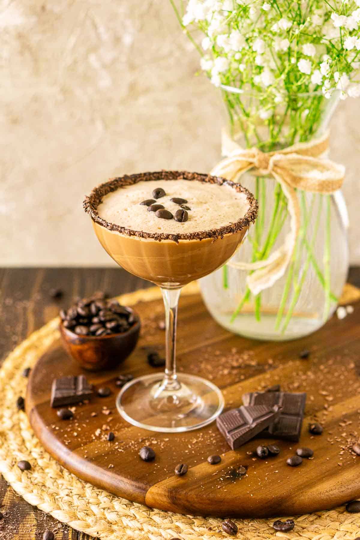A mocha martini on a wooden tray with pieces of chocolate and coffee beans scattered around it.