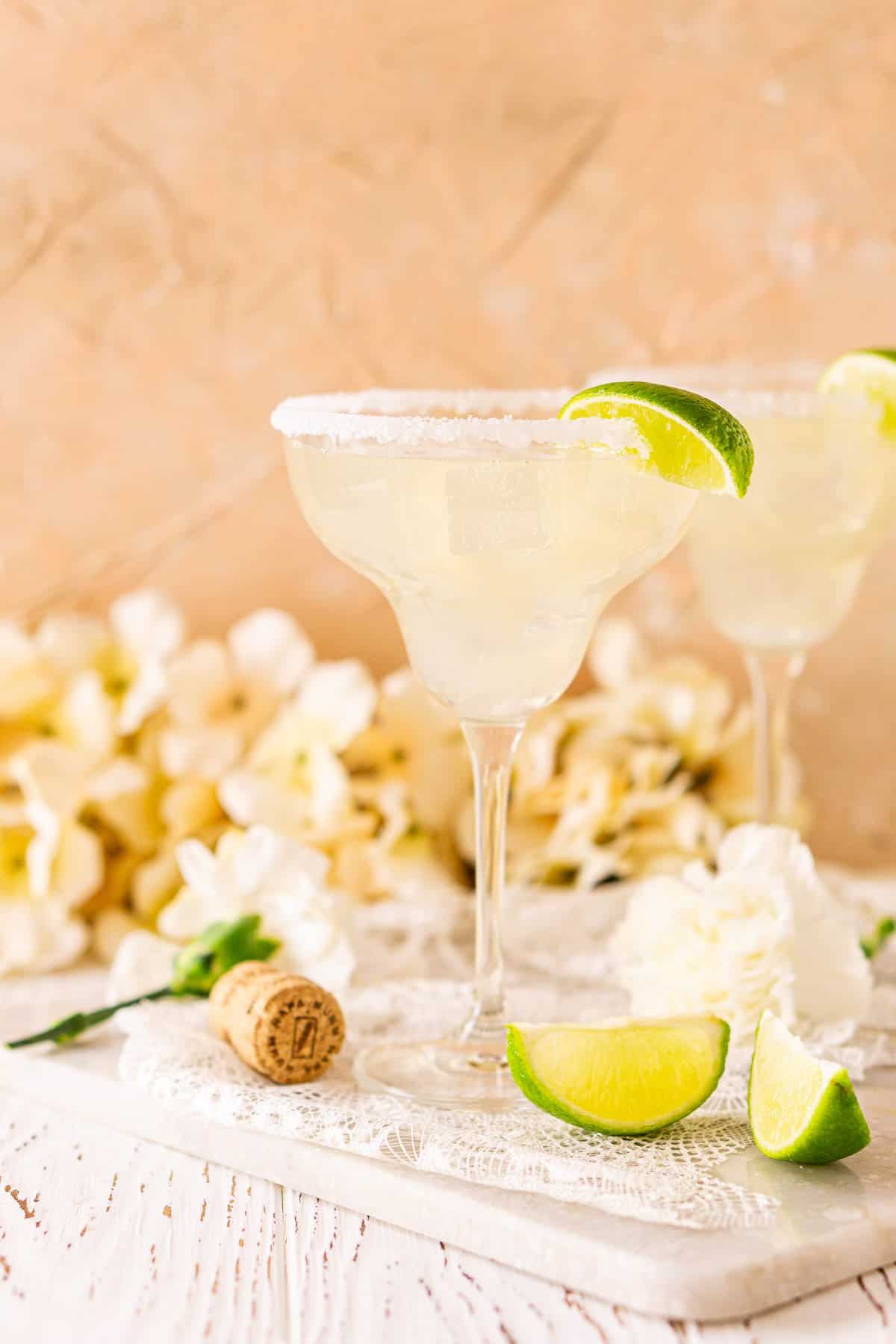 A straight-on view of the prosecco margarita on a white marble tray against a peach background with white flowers in the background and lime slices to the side.