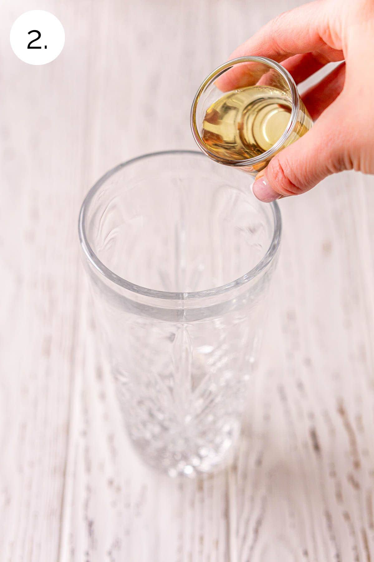A hand pouring a shot glass of tequila into a crystal cocktail shaker sitting on a white wooden surface.