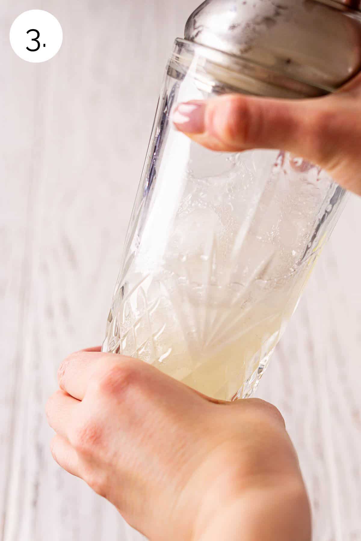 Two hands shaking the crystal cocktail shaker over a white wooden background to mix the drink.