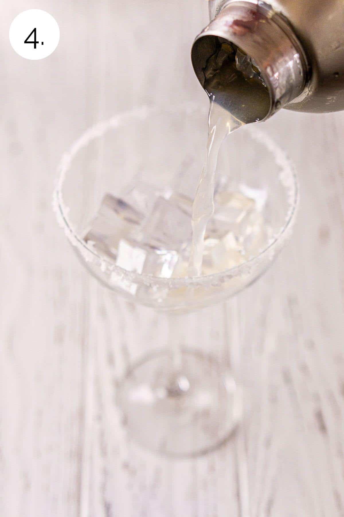 A cocktail shaker straining the margarita into a cocktail glass filled with ice on a white wooden surface.