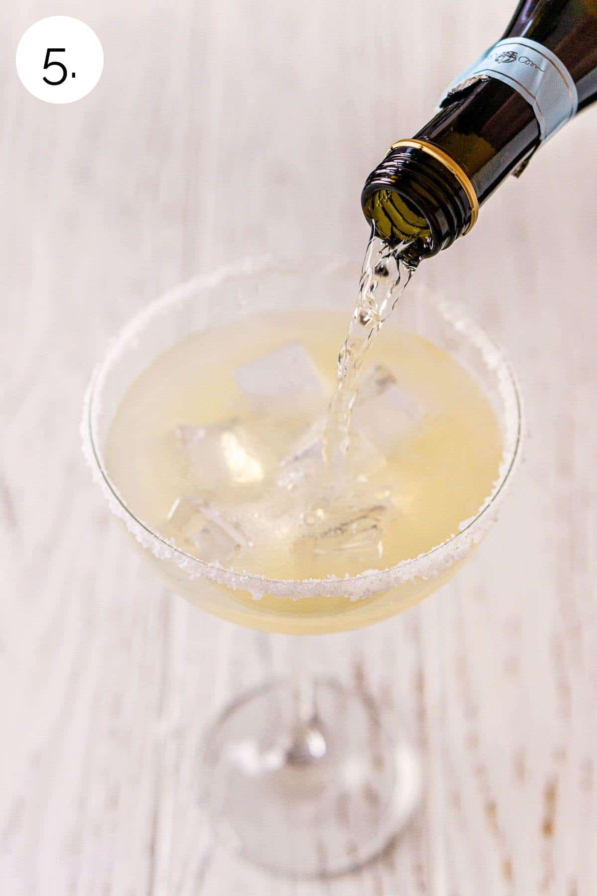 A small prosecco bottle pouring sparkling wine into the margarita glass after straining on a white wooden surface.