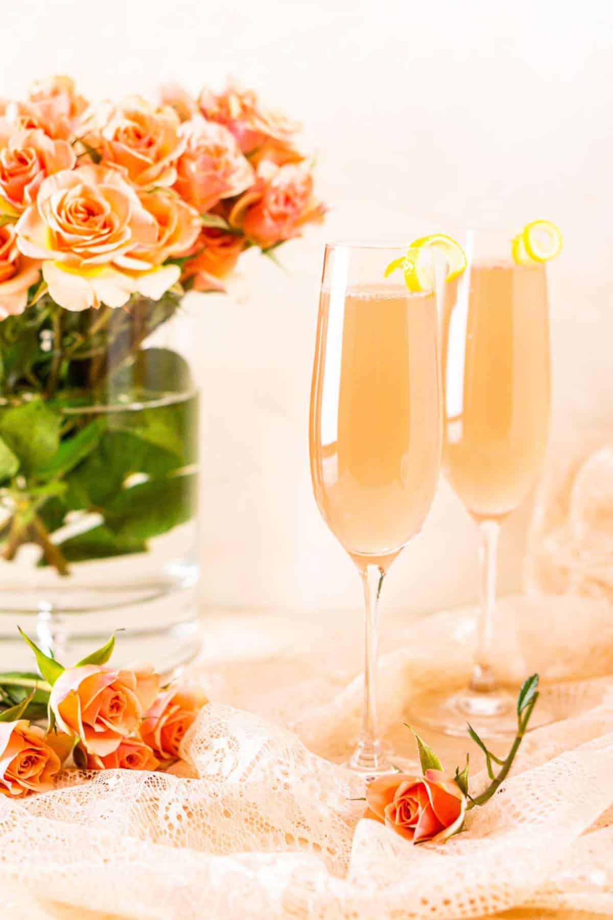 A sparkling rosé French 75 on pink lace with pink roses scattered around it and a flower vase in the background.