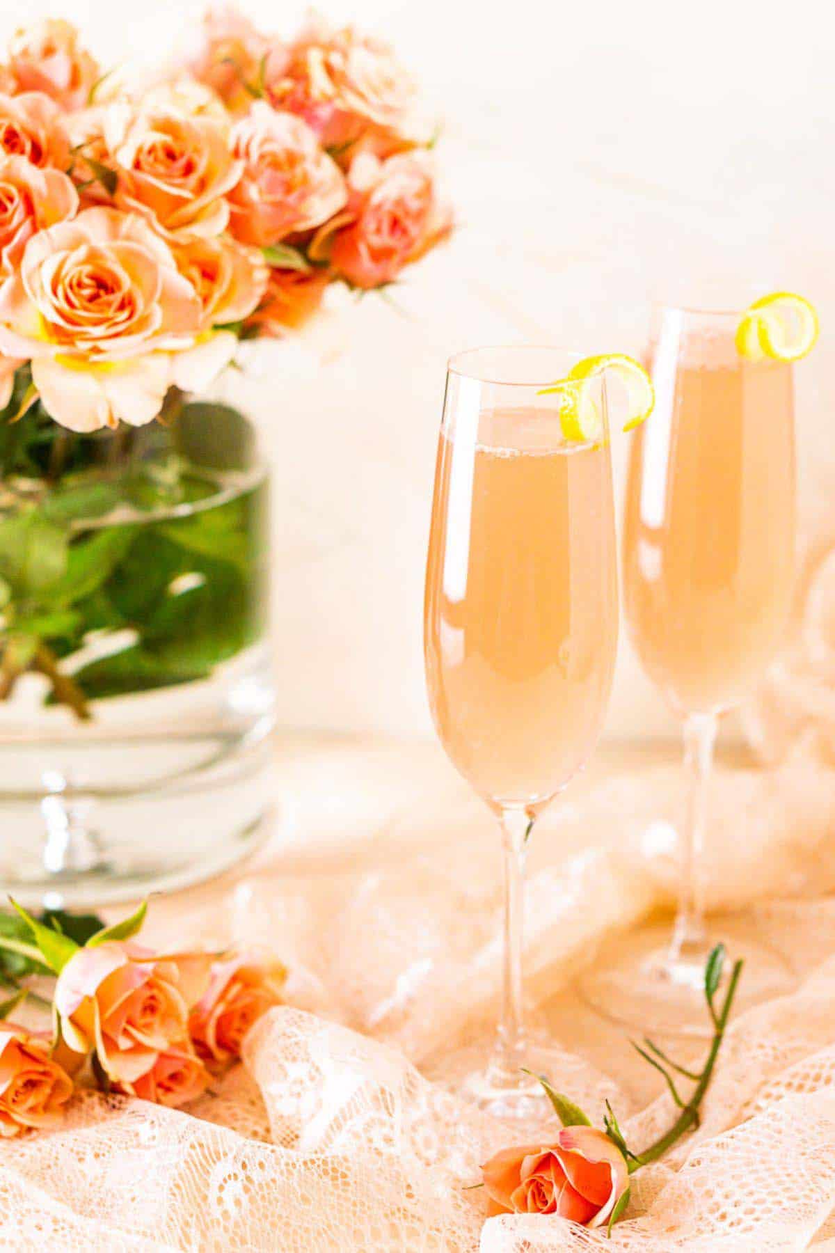 Two sparkling rosé French 75 cocktails with lemon twist on the side of the glass with pink roses in the background.