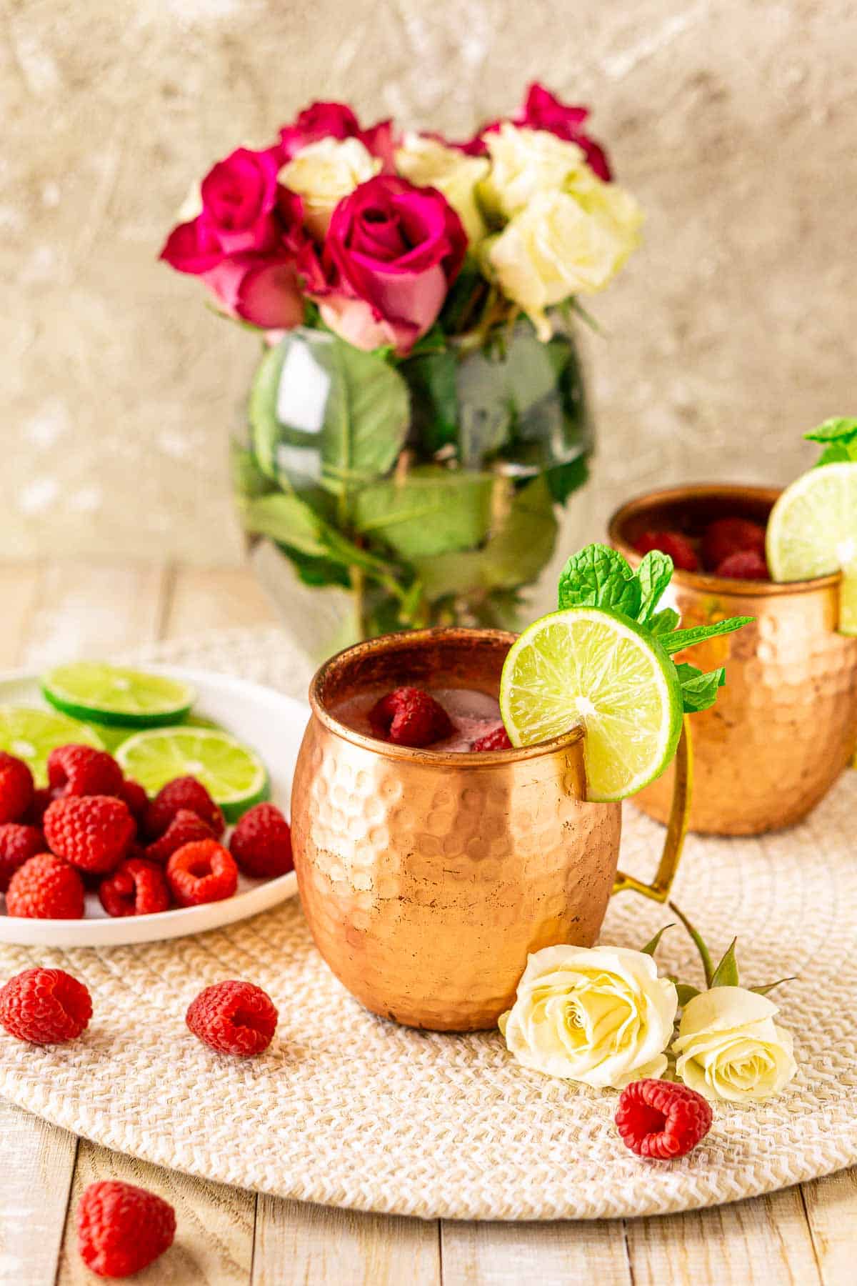Two raspberry Moscow Mule cocktails with flowers in the background and fresh berries scattered around the drinks.