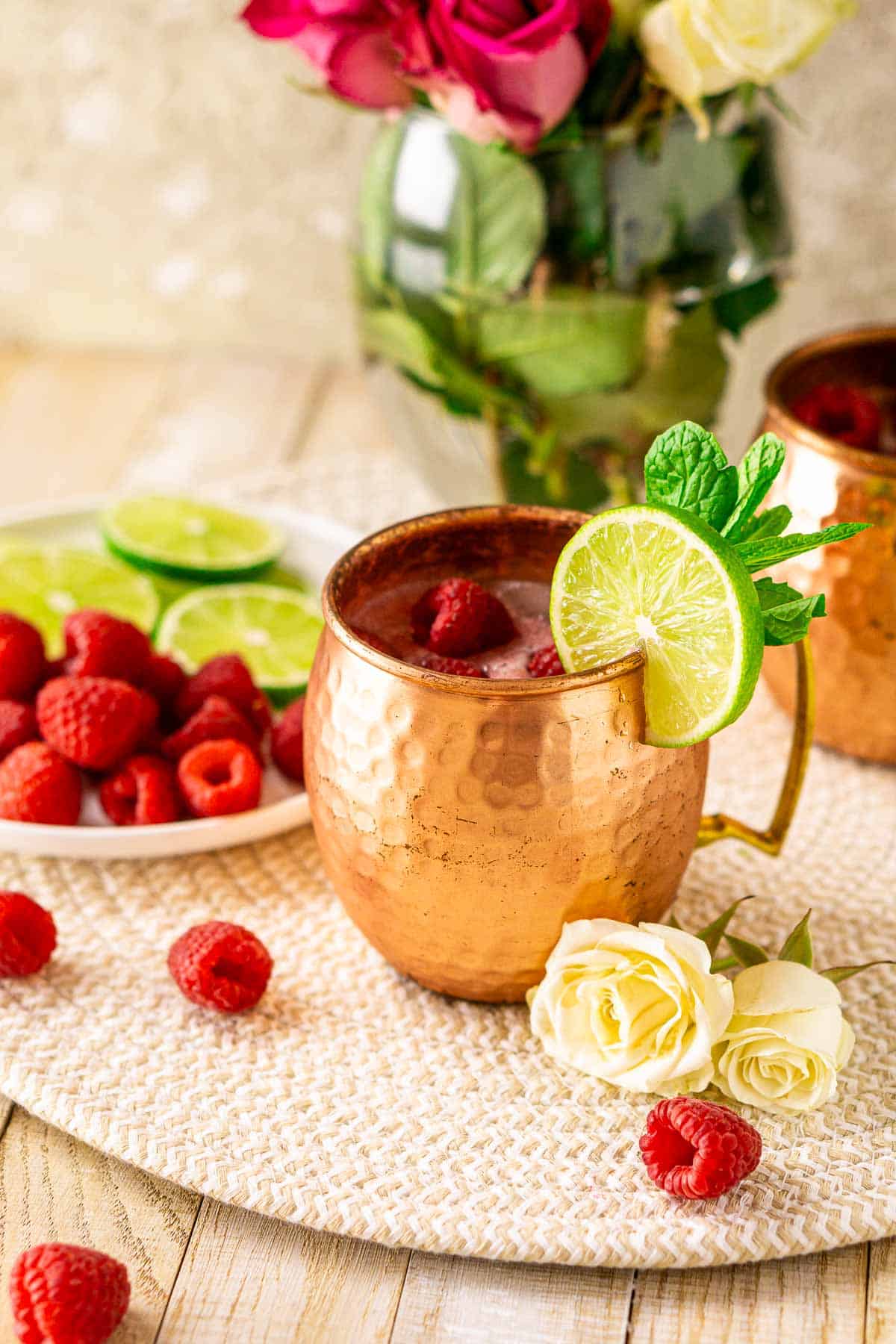 The raspberry Moscow mule on a straw placemat with a plate of raspberries to the side and a bouquet of flowers in the background.