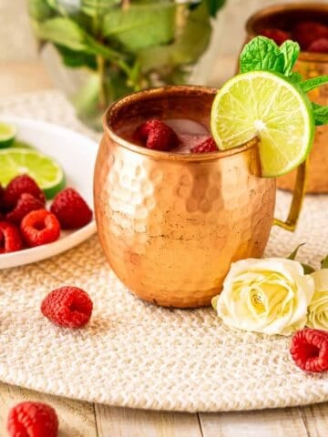 A raspberry Moscow mule on a cream-colored straw placemat with a white plate full of lime slices and raspberries to the left.