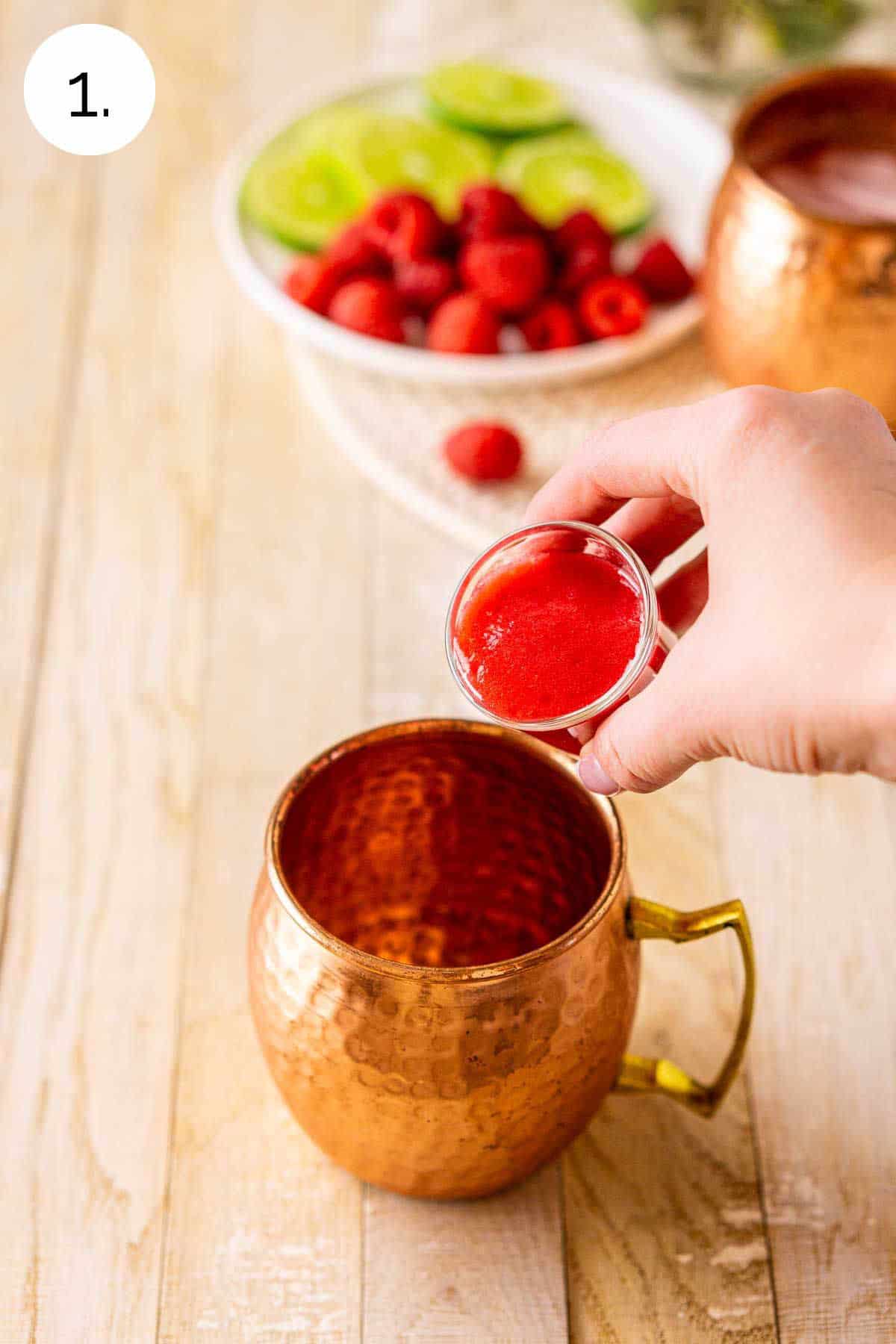 A hand pouring the raspberry purée into a copper mug on a cream-colored wooden board to start mixing the drink.