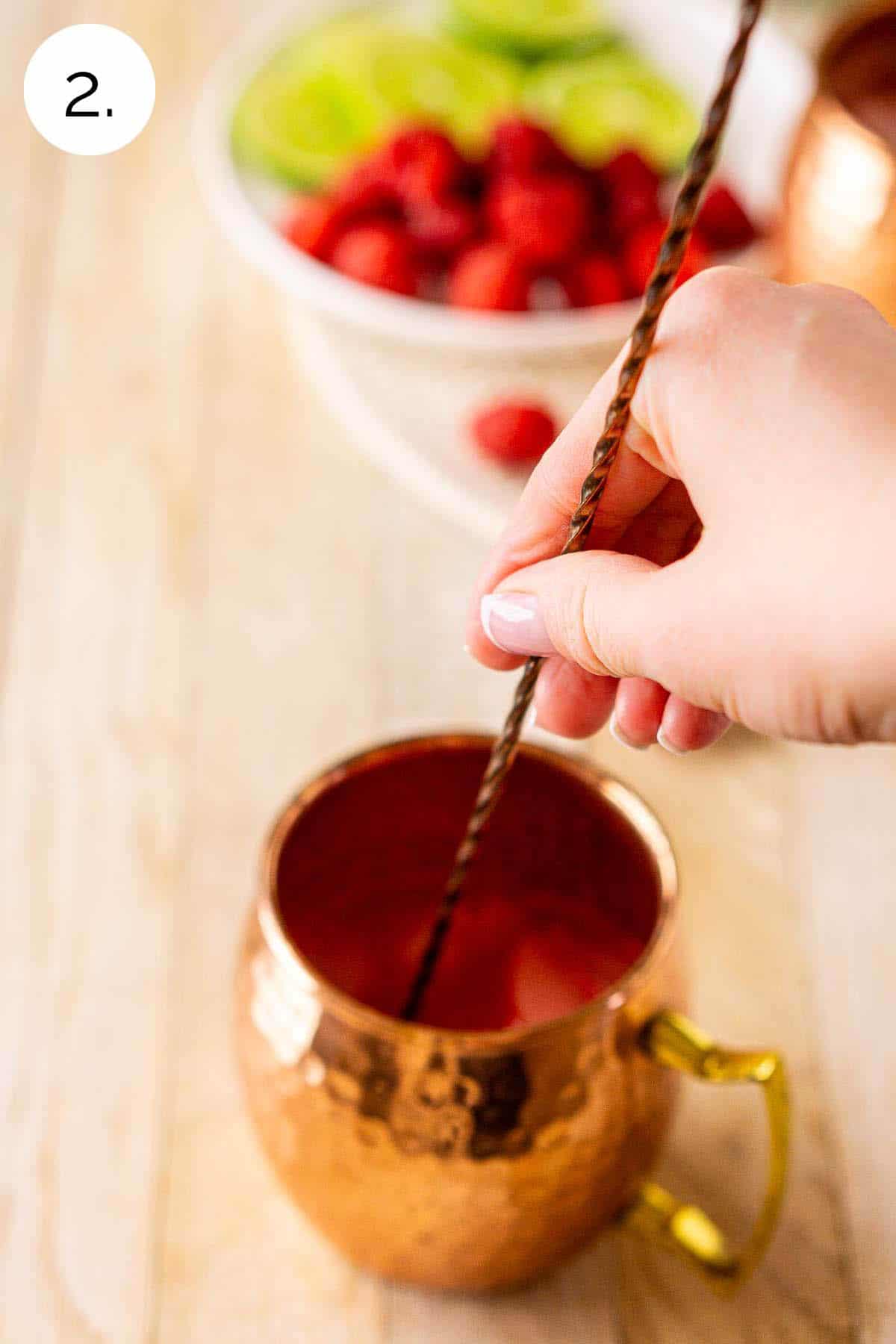 A hand stirring a long bar spoon to combine the raspberry mixture in the copper mug.