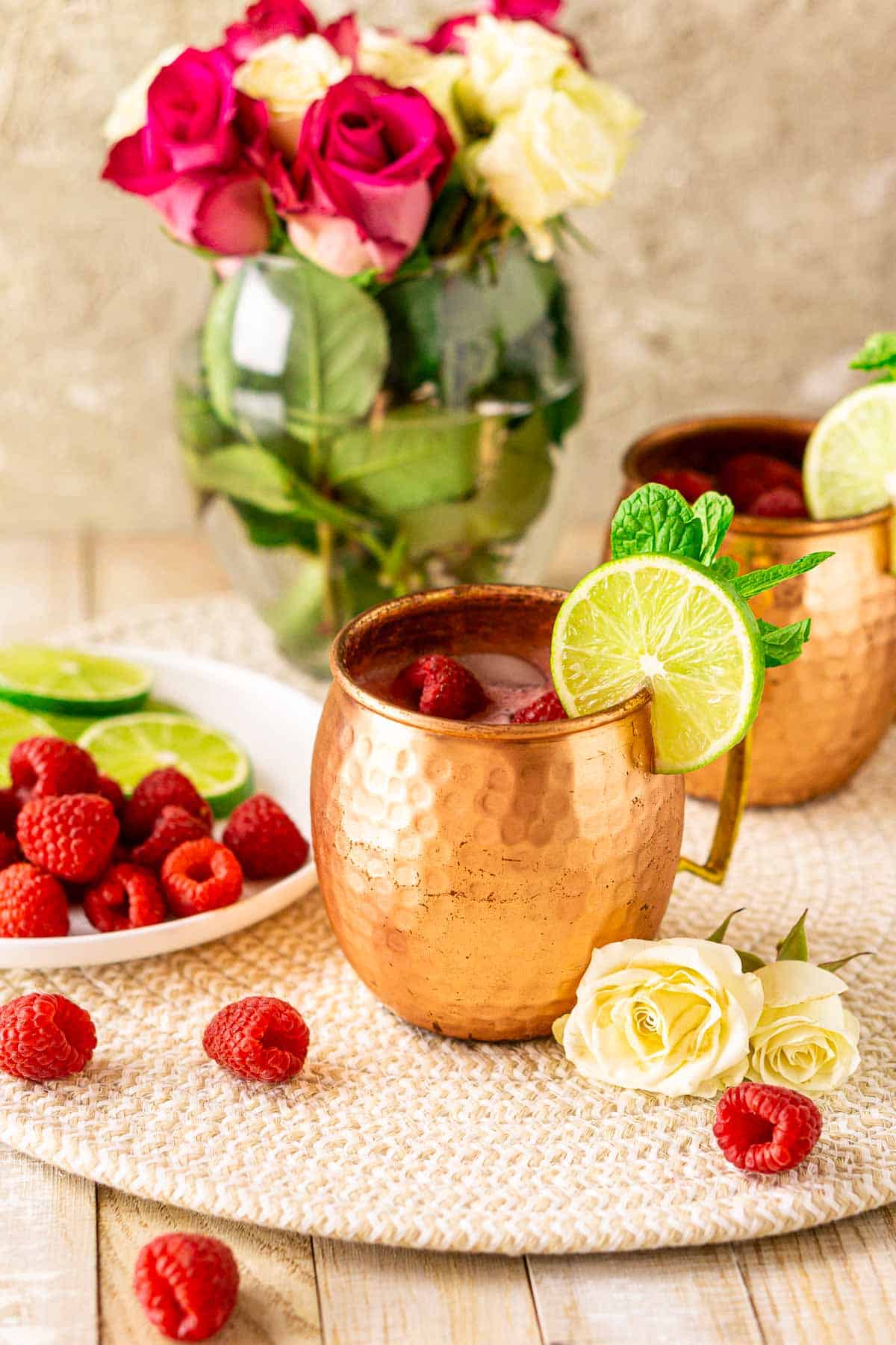 A raspberry Moscow mule with a white plate full of lime slices and raspberries to the left and pink and white flowers in the background.