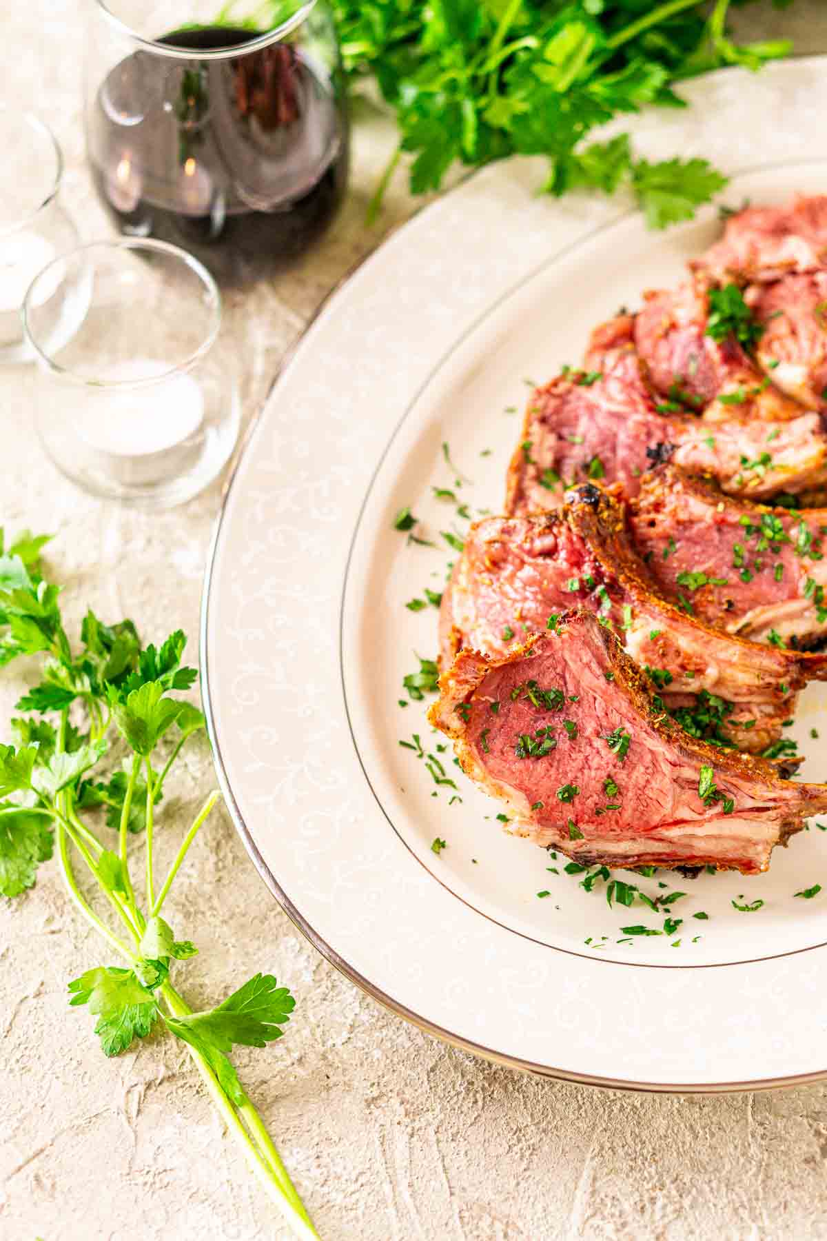 The smoked rack of lamb on a white serving platter with chopped parsley on top of a glass of wine and two candles to the side.