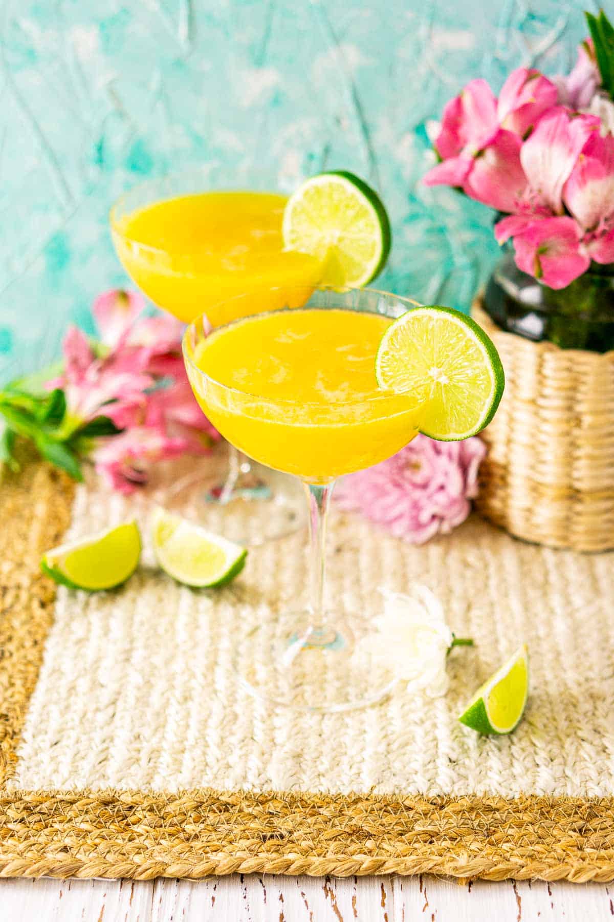 Two passion fruit daiquiri cocktails on a straw placemat with pink flowers to the right and lime slices around the drinks.