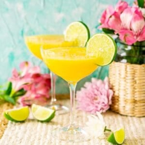 A passion fruit daiquiri on a straw placemat against a blue background with limes and colorful flowers around it.